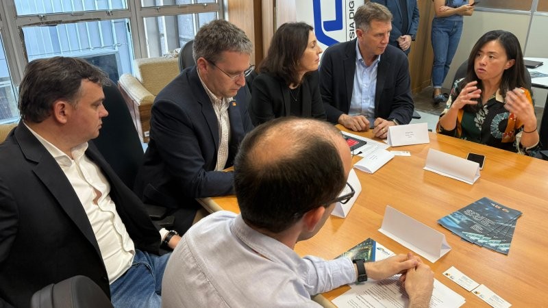 Grupo de pessoas em torno de uma mesa de reunião observa uma das integrantes da reunião, uma mulher gesticulando