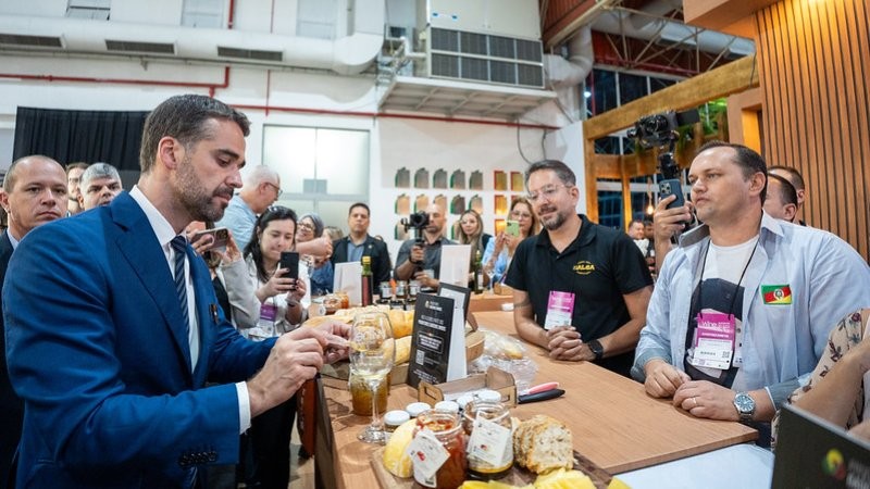Em uma mesa estão dispostos diferentes produtos como pães e geleias e azeites, enquanto um homem de terno, observado por outras pessoas ao entorno experimenta um quitute