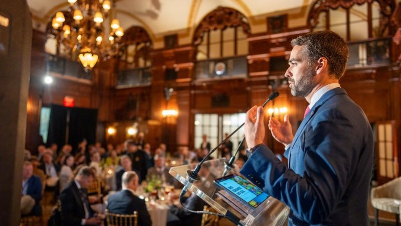 Um homem, à direita da foto, fala atrás de um púlpito para uma plateia num salão clássico