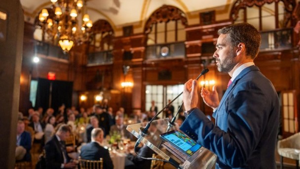 Um homem, à direita da foto, fala atrás de um púlpito para uma plateia num salão clássico 
