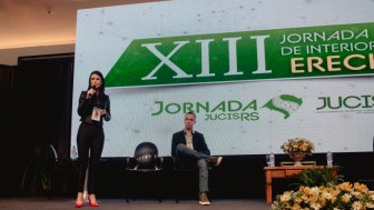 Uma mulher fala ao microfone em cima de um palco sendo observado por um homem sentado ao fundo do palco