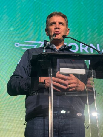 Foto de um homem branco, de meia idade, usando calça jeans e jaqueta azul escura. Ele está de pé em um palco, falando ao microfone. Ao fundo, uma tela verde.
