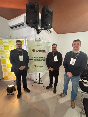 Imagem de três homens brancos, de pé, com um banner escrito "badesul cidades" no centro