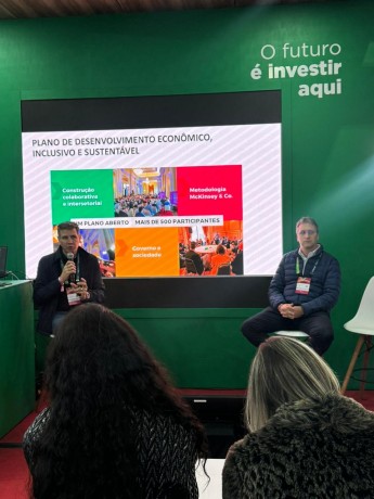 dois homens brancos sentados. Um deles está segurando um microfone. Atrás, se vê um slide escrito "Plano de Desenvolvimento Econômico, Inclusivo e Sustentável"