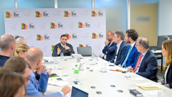 foto mostra uma mesa de reuniões e um grupo de pessoas em torno da mesa conversando