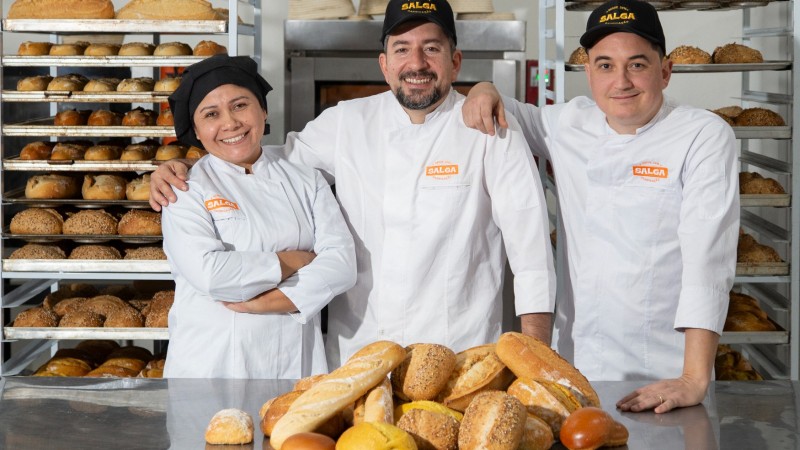 Foto de três pessoas, dois homens e uma mulher, em uma fábrica de pães.