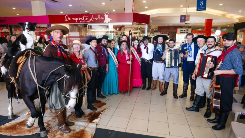 Grupo de pessoas, homens e mulheres, perfilados lado a lado, vestindo trajes tipicamente ga&uacute;chos, incluindo um cavalo com um cachorro em cima, posam para a foto dentro de um shopping