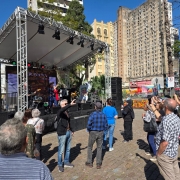 Músicos se apresentam num palco externo, pessoas observam e tiram fotos com os celulares