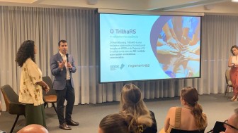 Um homem e uma mulher em um palco, em frente a um telão onde se lê "O Trilha RS"