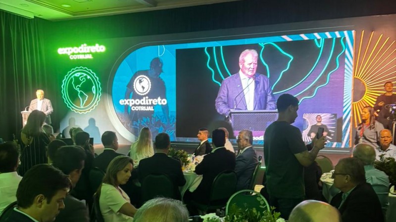 Foto de um grande sal&atilde;o com pessoas sentadas em torno de mesas, ao fundo um palco uma pessoa fala atr&aacute;s de um p&uacute;lpito e sua imagem &eacute; projetada no tel&atilde;o