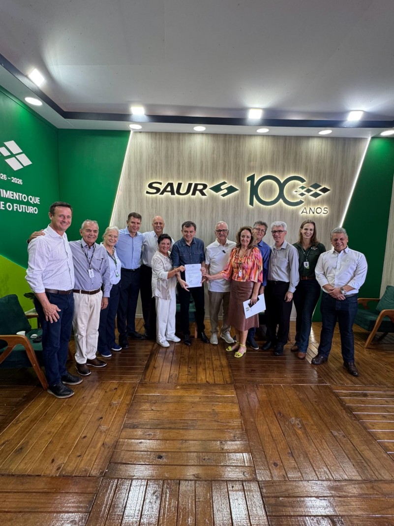 Um grupo de pessoas est&aacute; de p&eacute;, posando paraa foto. Ao fundo ,se v&ecirc; escrito Saur 100 anos