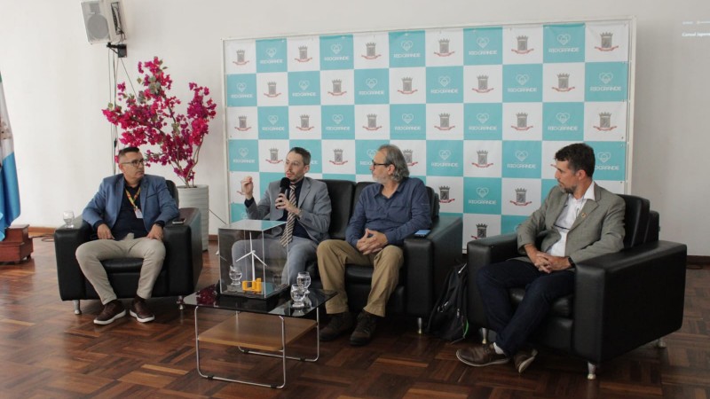 foto de quatro homens sentados em um sof&aacute;. Um deles fala ao microfone. H&aacute; um painel quadriculado branco e azul ao fundo 