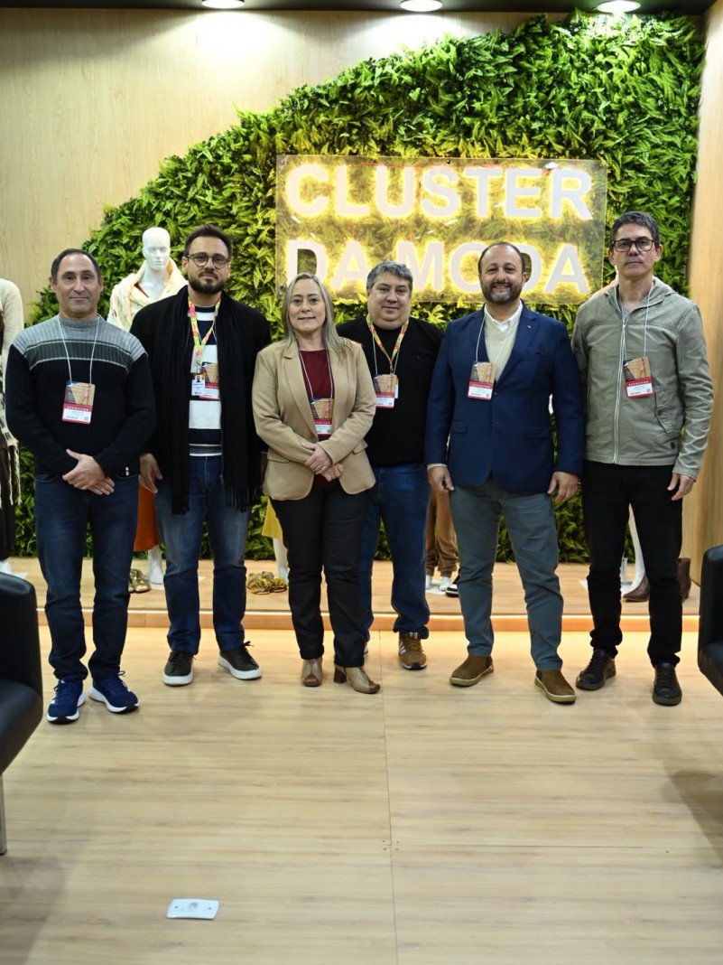 Foto de um grupo de pessoas de p&eacute;, lado a lado. Atr&aacute;s h&aacute; plantas verdes e um letreiro neon onde se l&ecirc; "cluster da moda"
