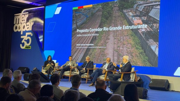 Foto de cinco pessoas sentadas em cadeiras em um palco, em um painel de debate sobre ferrovias